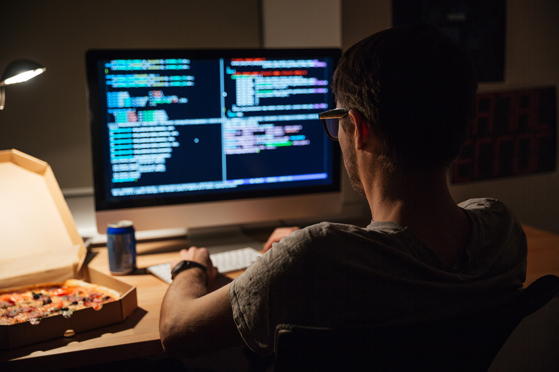 Back view of focused young programmer in glasses writing code and eating pizza